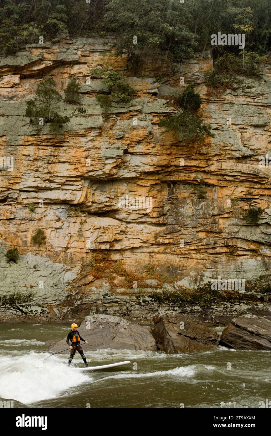 Uomo in piedi per il paddleboarder che fa surf su un'onda su un fiume sotto una grande scogliera sotto la pioggia. Foto Stock