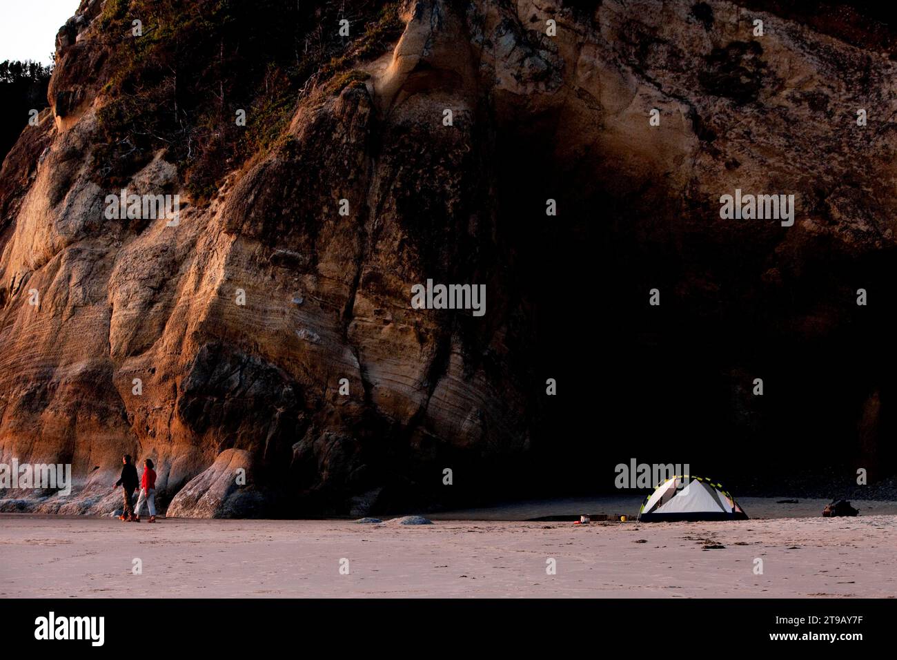 Coppia di uomini e donne che camminano sulla spiaggia sotto le scogliere accanto alla loro tenda al tramonto. Foto Stock