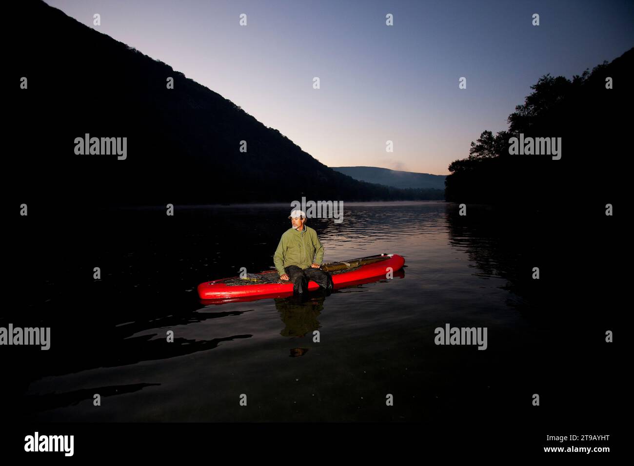 Un uomo seduto su una tavola da paddle stand-up prima dell'alba con un fucile. Foto Stock