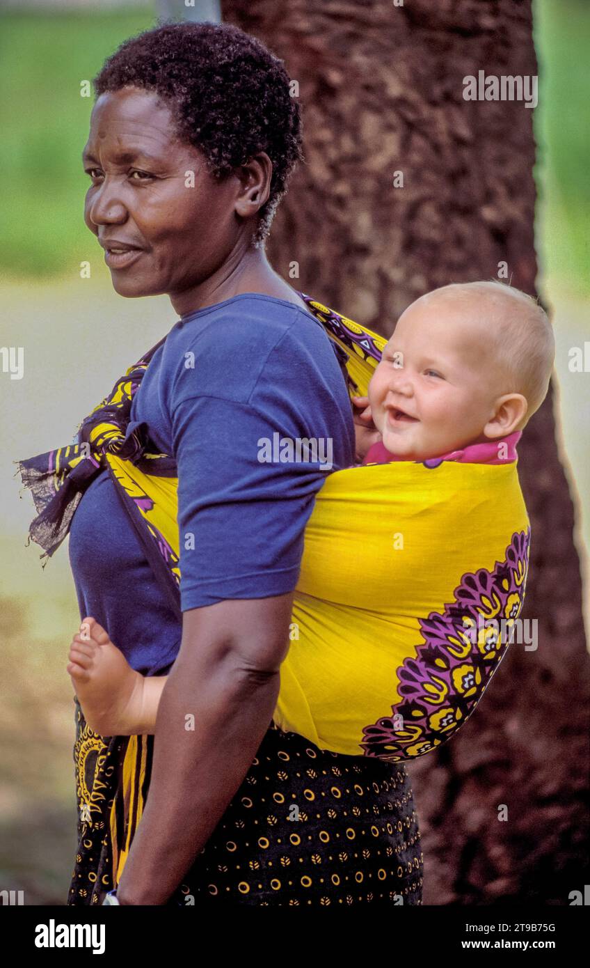 Tanzania, Una tata nera sta portando un bambino bianco sulla schiena. Foto Stock