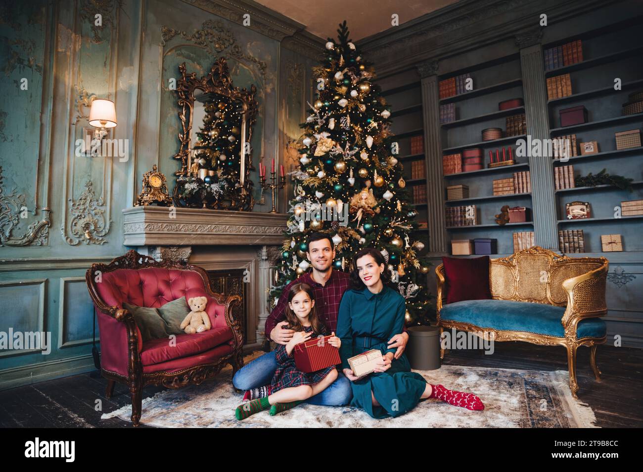Famiglia con un bambino che siede in una camera in stile vintage con un albero di Natale riccamente decorato e che regala regali Foto Stock