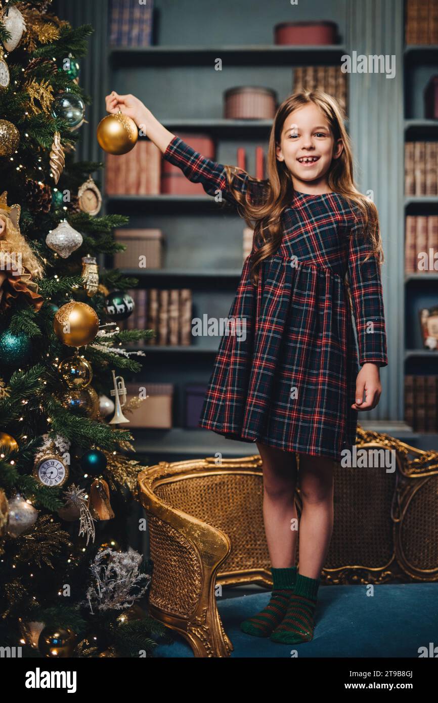 Gioiosa ragazza con un abito tartan che regge un ornamento natalizio dorato, con un albero riccamente decorato e una camera elegante Foto Stock