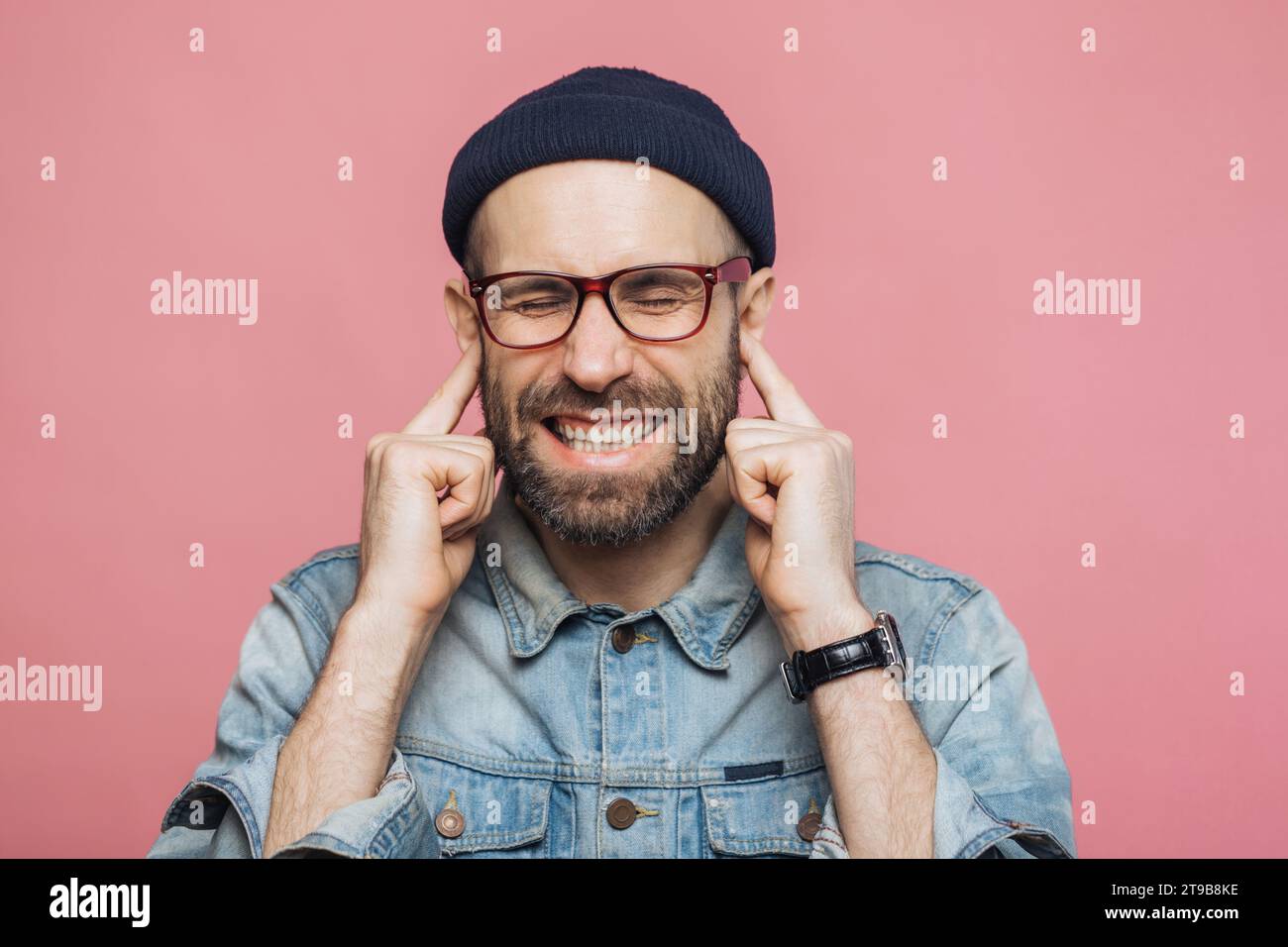 Uomo con berretto e occhiali che stringe le mani sulle orecchie, vestito in denim, su uno sfondo rosa Foto Stock