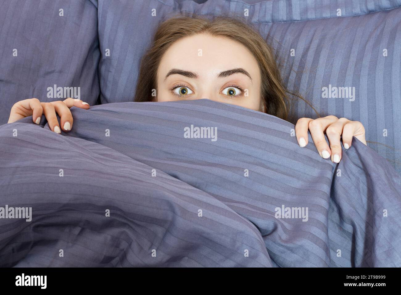 La ragazza adolescente con gli occhi aperti si copre la faccia con una coperta blu la mattina. Bella donna sdraiata nel letto e addormentata. Non dormi abbastanza Foto Stock