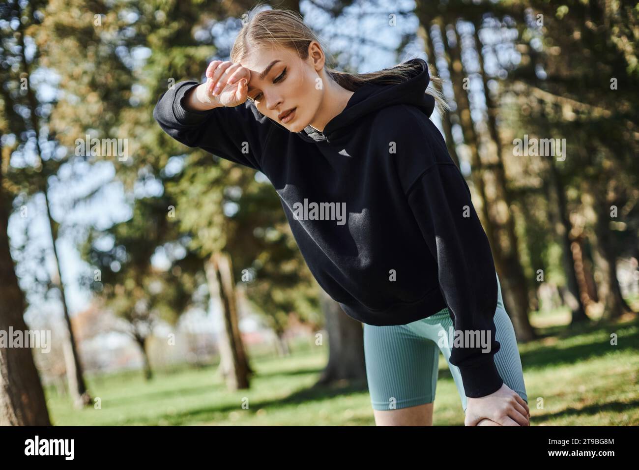 giovane donna stanca con i capelli biondi e abbigliamento sportivo che pulisce il sudore dopo aver lavorato nel parco verde Foto Stock