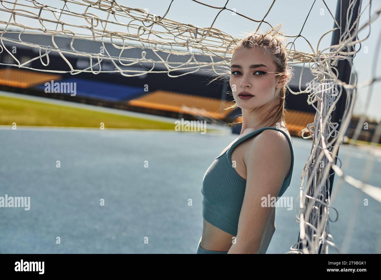 donna sportiva bionda con coda di cavallo in piedi vicino alla rete dopo l'allenamento nello stadio, fitness urbano Foto Stock