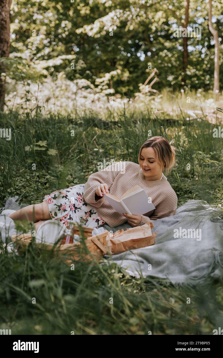 Donna sorridente sdraiata su una coperta e leggendo un libro durante il giorno d'estate Foto Stock