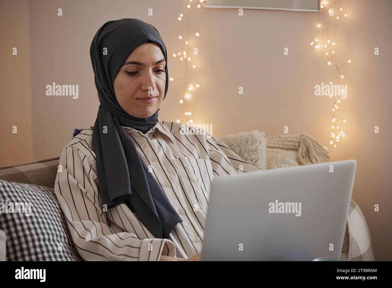 Giovane freelance che indossa l'hijab e lavora su un computer portatile in salotto Foto Stock