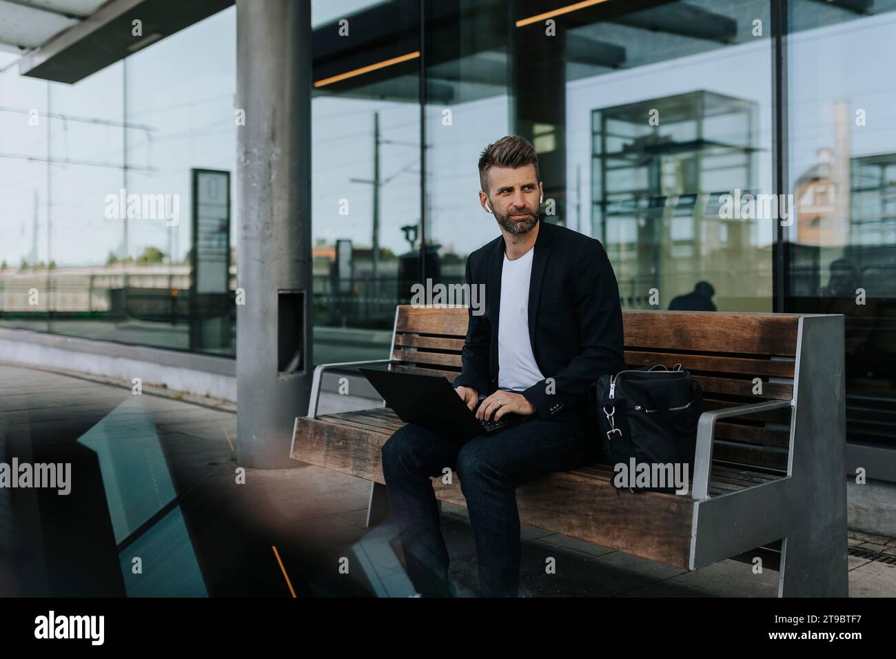 Uomo d'affari seduto con un computer portatile sulla panchina mentre aspetta alla stazione ferroviaria Foto Stock