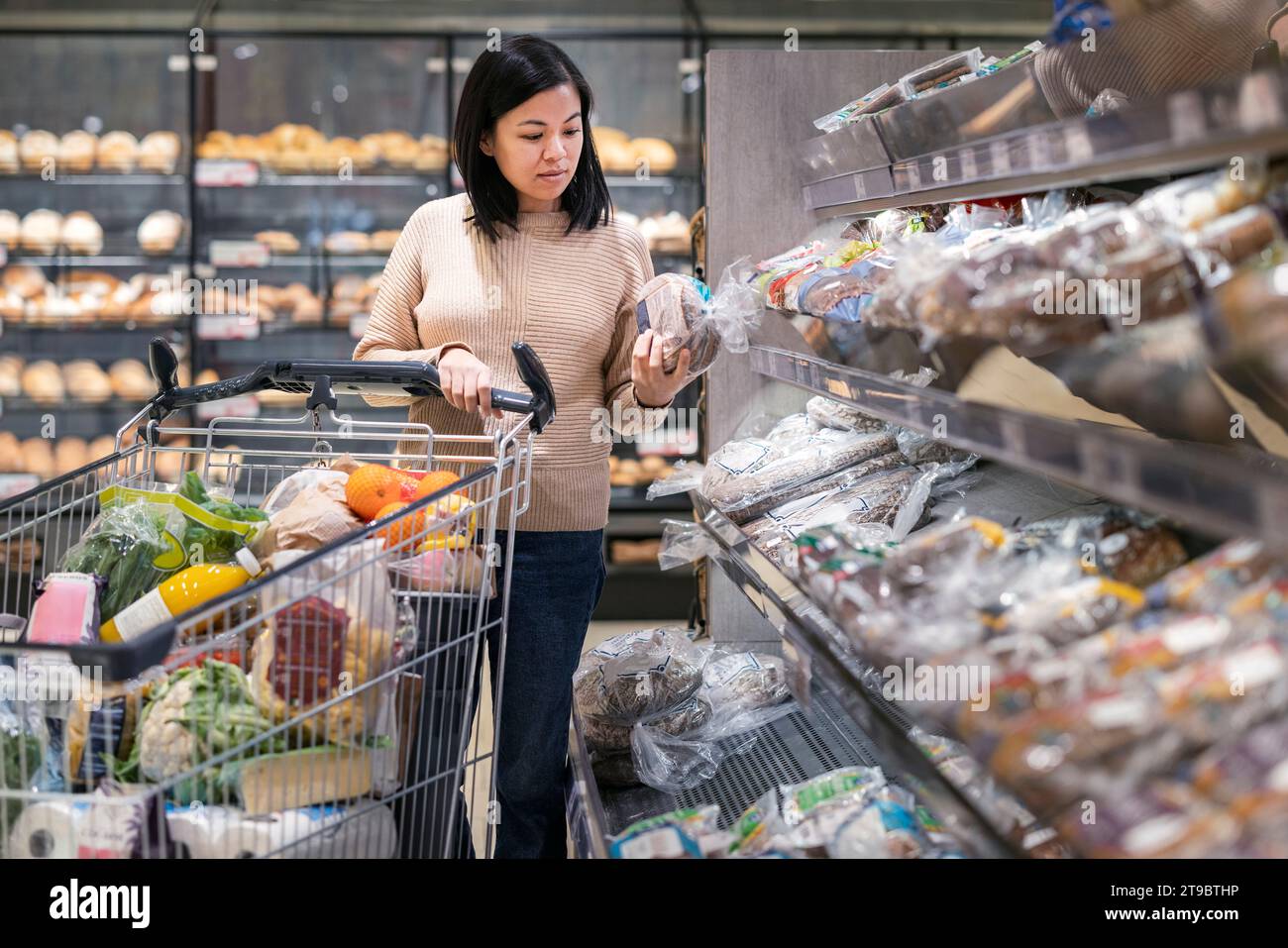 Giovane donna che legge l'etichetta sulla confezione del pane mentre compra generi alimentari al supermercato Foto Stock