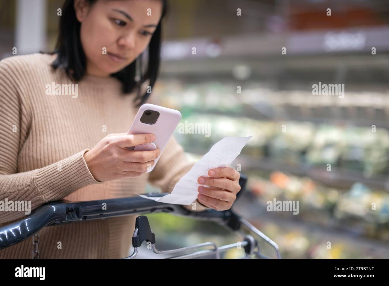 Giovane donna che esamina la ricevuta mentre usa lo smartphone al supermercato Foto Stock