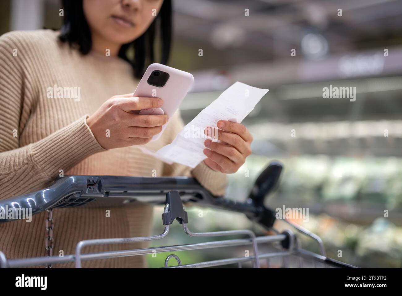 Giovane donna che sta esaminando la lista mentre usa lo smartphone al supermercato Foto Stock