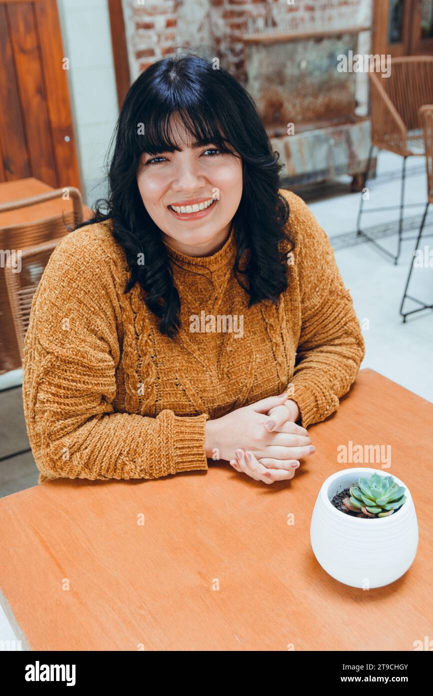 l'immagine verticale della giovane donna venezuelana latina, vestita di marrone, è seduta con le mani sul tavolo, guardando la macchina fotografica sorridendo felice attesa nel caffè Foto Stock l'immagine verticale della giovane donna venezuelana latina, vestita di marrone, è seduta con le mani sul tavolo, guardando la macchina fotografica sorridendo felice attesa nel caffè Foto Stock