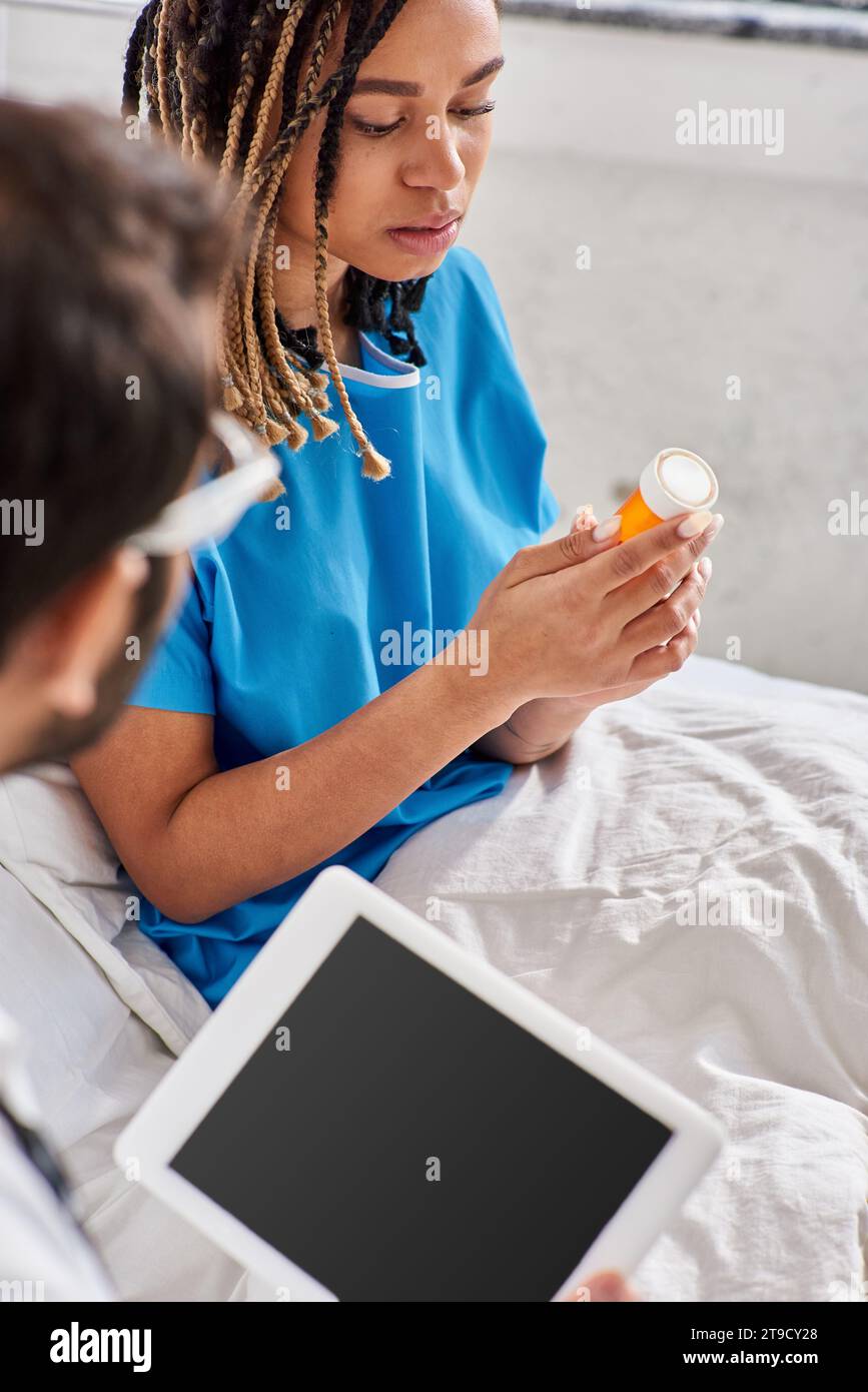 concentrati su una donna afroamericana malata seduta sul letto e guardando le sue pillole accanto al medico indiano Foto Stock