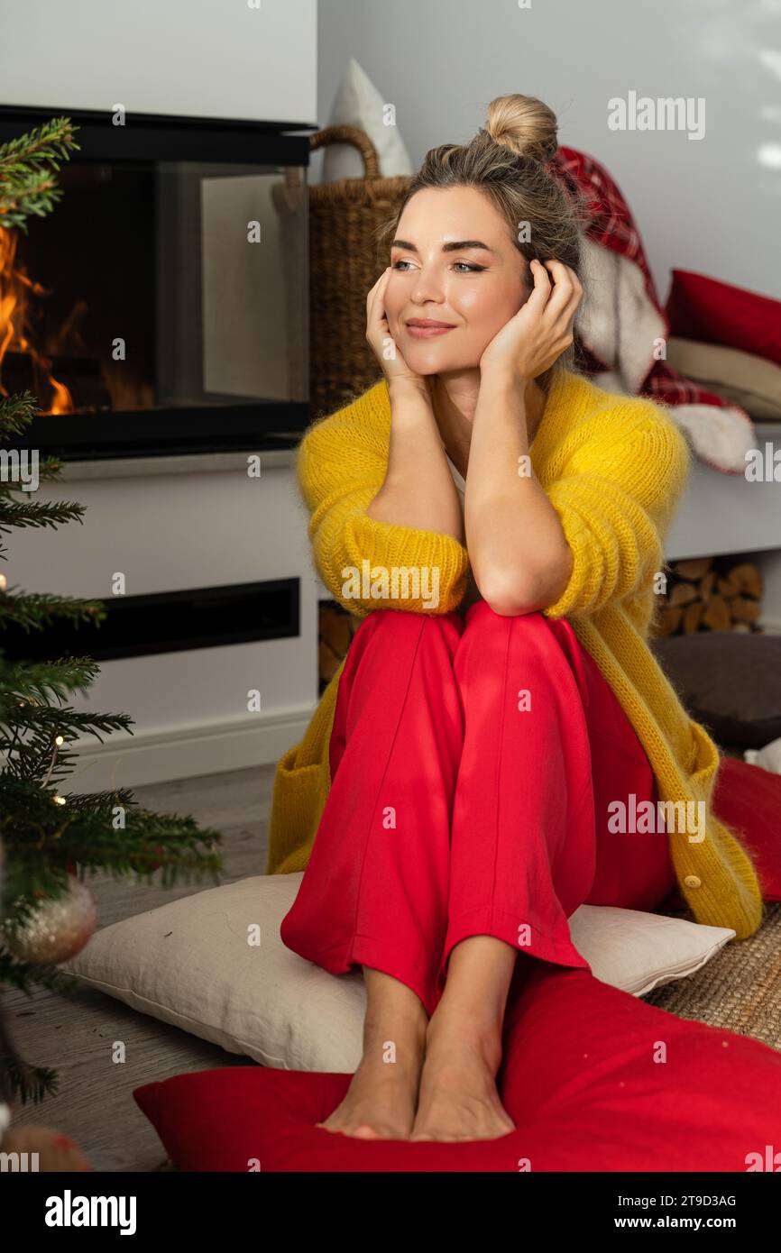 Giovane donna siede accanto a un caminetto luminoso in un accogliente soggiorno adornato con un albero di Natale e decorazioni festive. Foto Stock