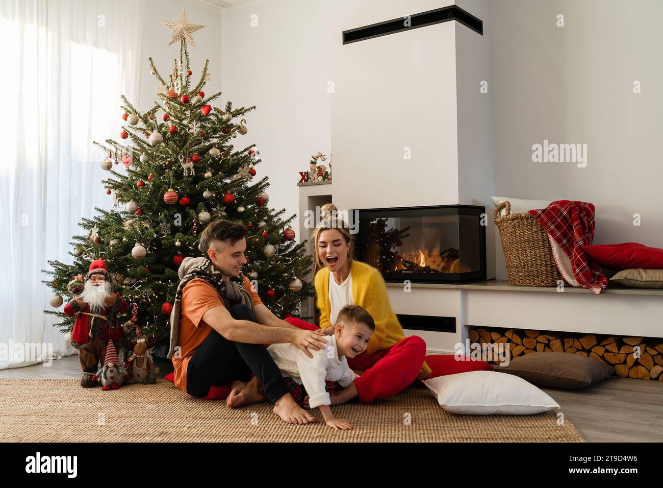 La giovane famiglia felice siede accanto a un caminetto luminoso in un accogliente soggiorno, adornato con un albero di Natale e decorazioni festive. Foto Stock