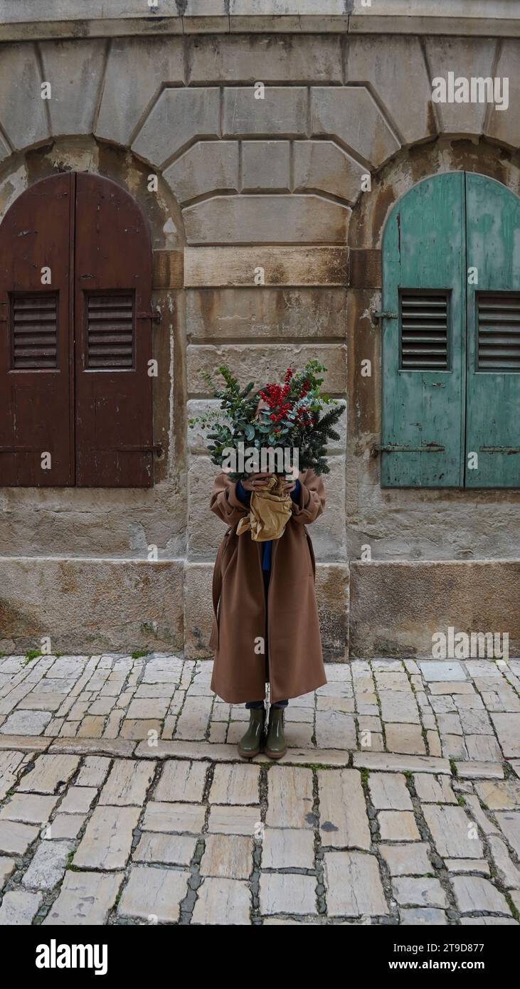 Una ragazza con un cappotto beige tiene un bouquet natalizio con rami di abete, bacche invernali, eucalipto sullo sfondo di una casa in pietra Foto Stock