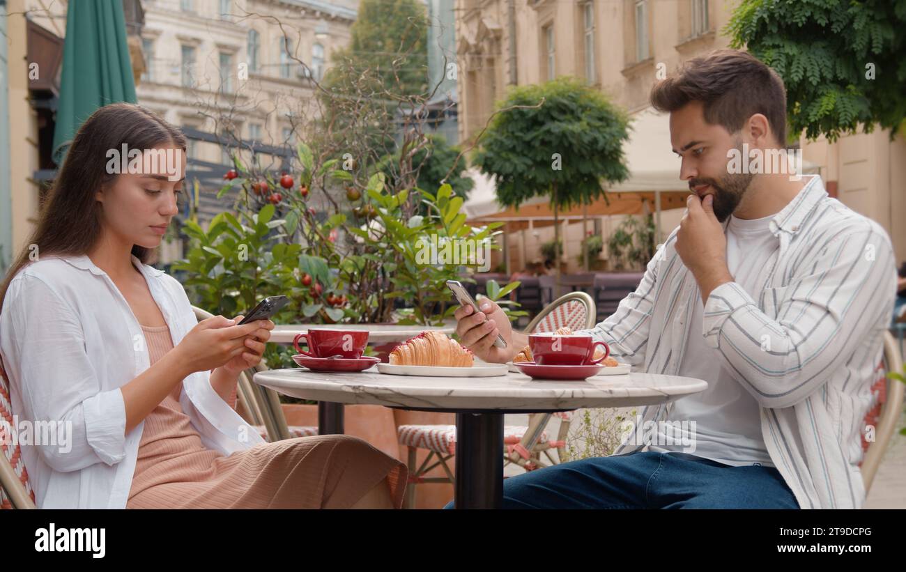 Coppia caucasica nel caffè della città che usa il telefono cellulare che invia messaggi al lavoro uomo serio donna che fa colazione fuori insieme gen z bevendo caffè chiacchierando Foto Stock