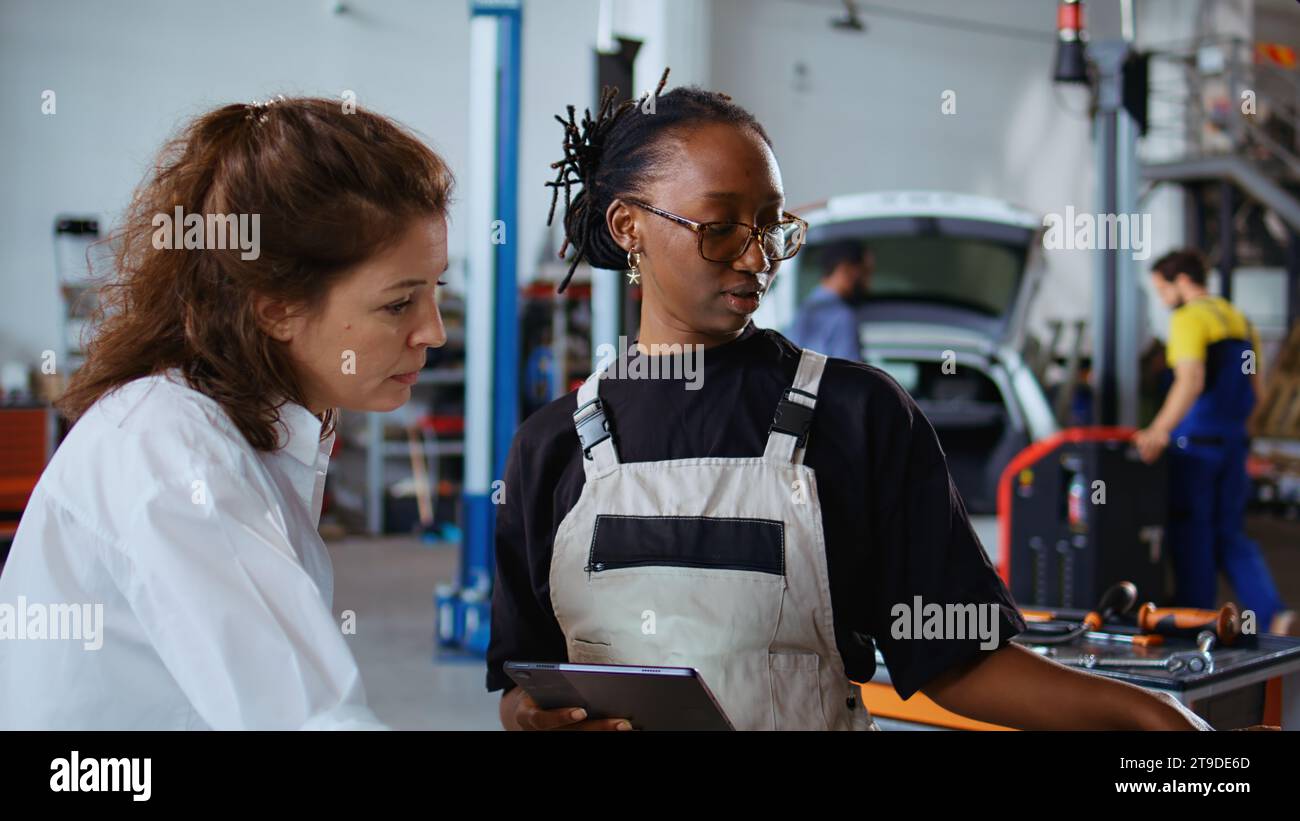 I lavoratori delle officine di riparazione si scambiano idee sui modi per riparare l'auto, utilizzando tablet e notebook. Colleghi che eseguono interventi di manutenzione sul veicolo guasto, controllano i componenti difettosi utilizzando dispositivi elettronici, zoom indietro Foto Stock