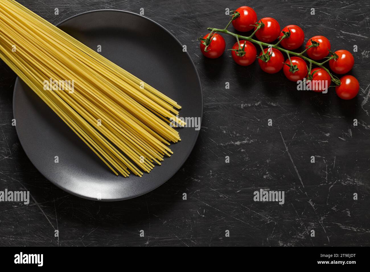 Spaghetti di pasta cruda su piatto grigio e ciuffo di pomodori ciliegini vista dall'alto sfondo scuro, con spazio per copiare il testo. Foto Stock