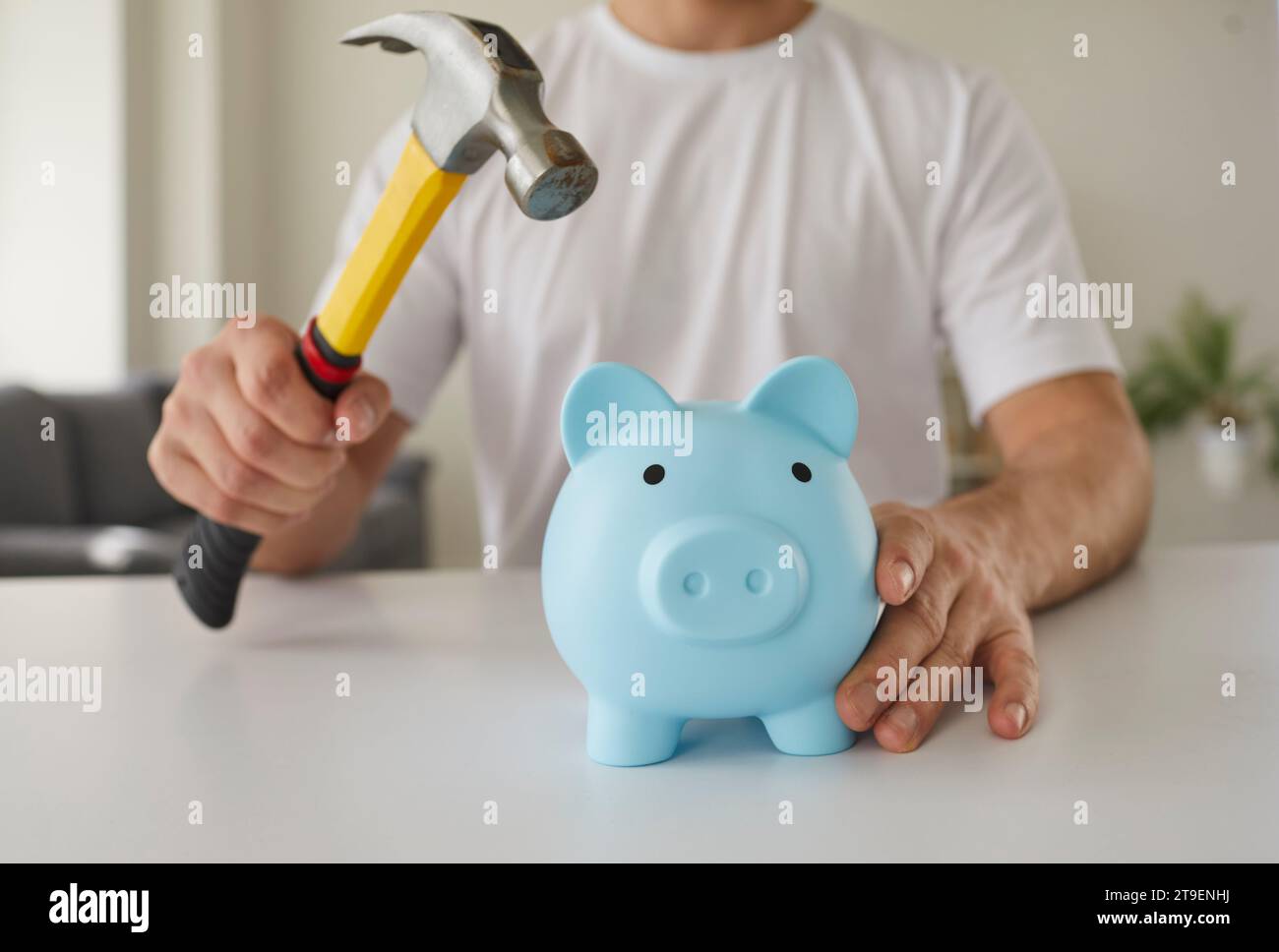 Il giovane tiene HAMMER e vuole rompere il salvadanaio blu con i suoi risparmi di denaro Foto Stock