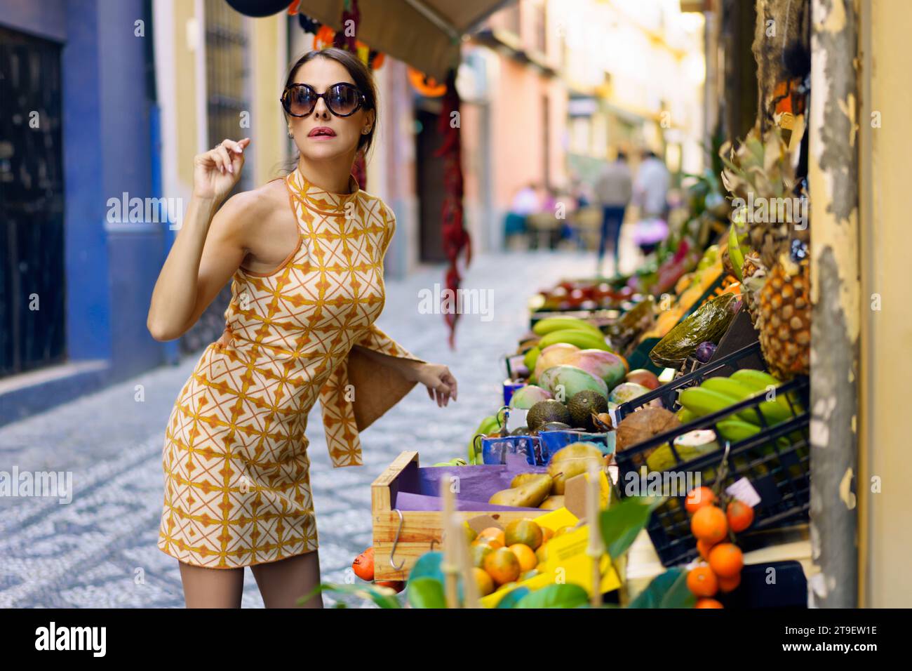 Donna elegante e positiva in abito alla moda, con trucco e occhiali da vista in piedi e inclinati in avanti da frutta fresca selezionata in vassoi e cesti in città Foto Stock