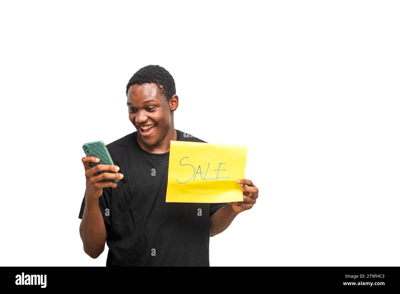 Giovane in possesso di cartellone pubblicitario giallo in vendita mentre utilizza lo smartphone. Eccitato uomo africano su camicia nera con faccia a sorpresa. Black Friday sal Foto Stock