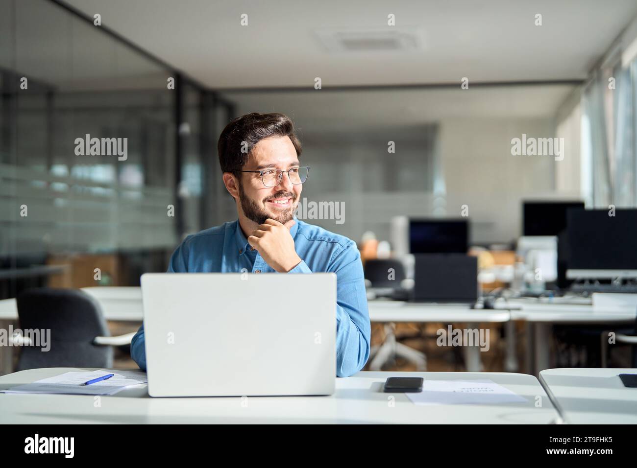 Felice giovane uomo d'affari che lavora con un computer portatile e guarda lontano dalla scrivania. Foto Stock