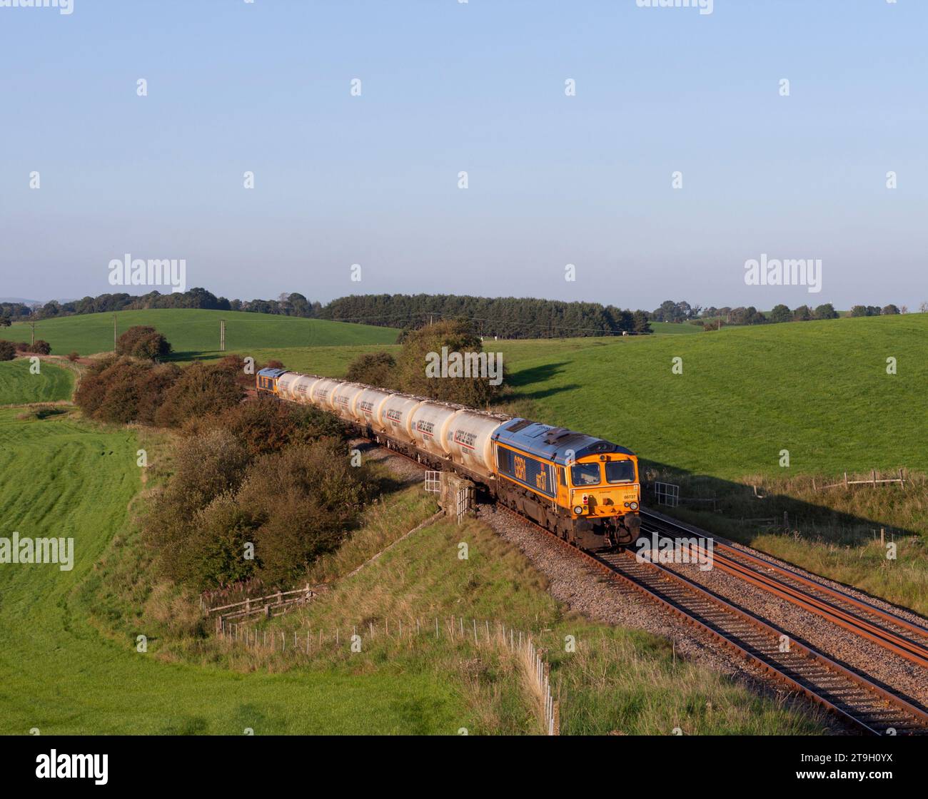 GB Railfreight classe 66 locomotiva che passa Newsholme sulla linea ferroviaria della Ribble Valley con un treno merci che trasporta cemento Foto Stock