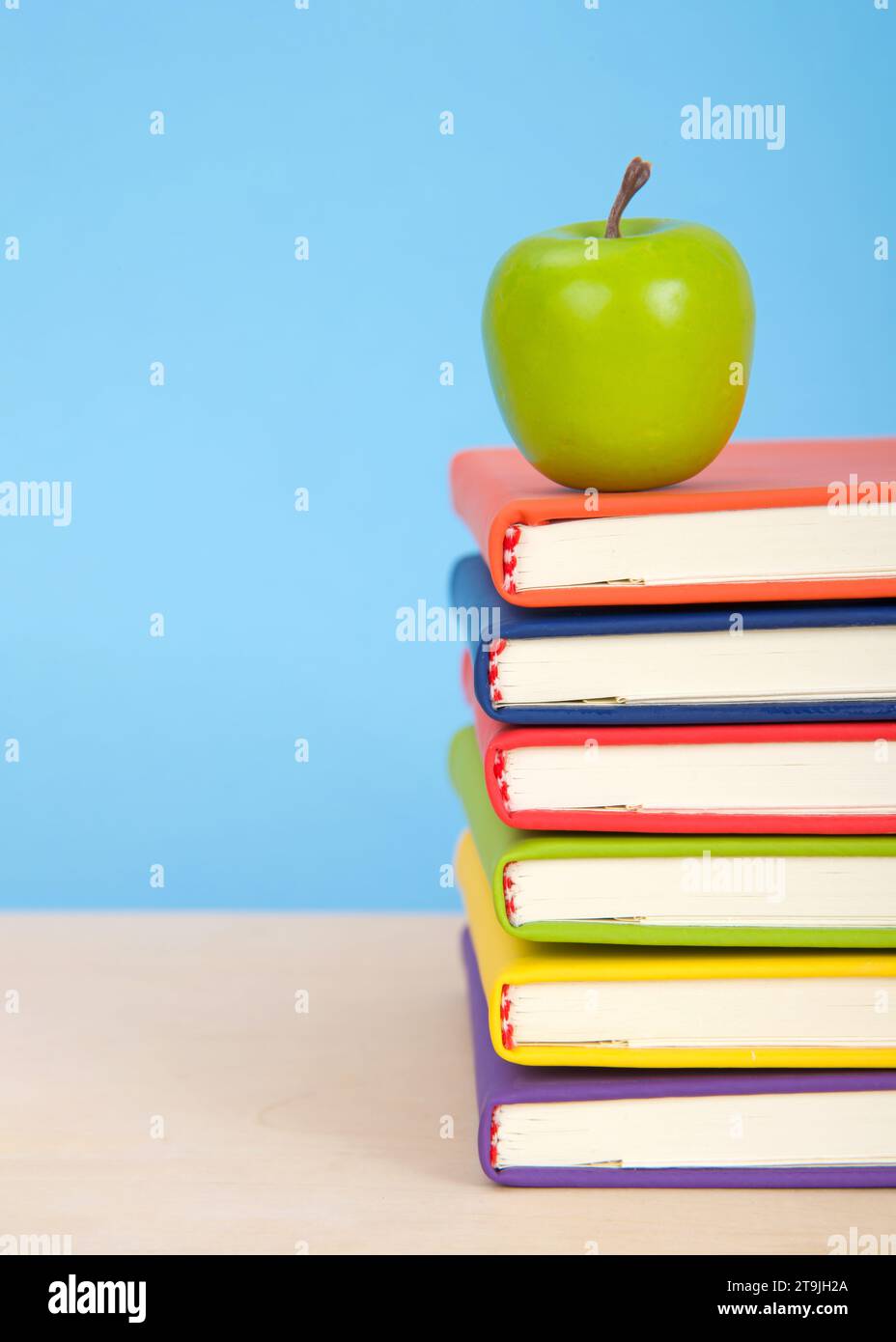 Libri rilegati colorati e luminosi impilati su un tavolo di legno chiaro con sfondo blu. Mela verde sopra i libri impilati. Foto Stock