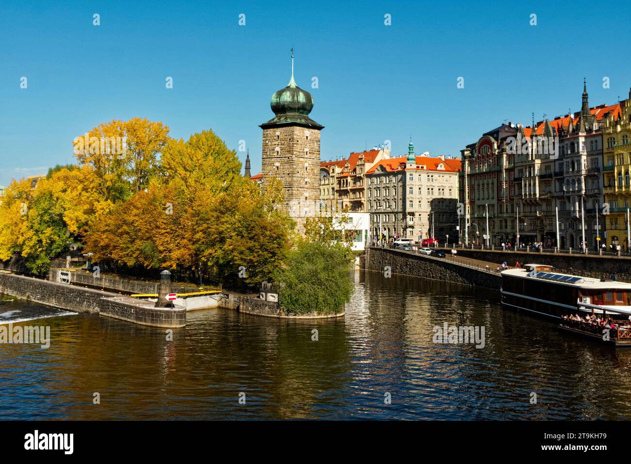 Una scena fluviale con una torre a cupola verde sulla riva sinistra e colorati edifici sulla destra. Antica torre sulla riva del fiume in autunno. Autunno Pragu Foto Stock