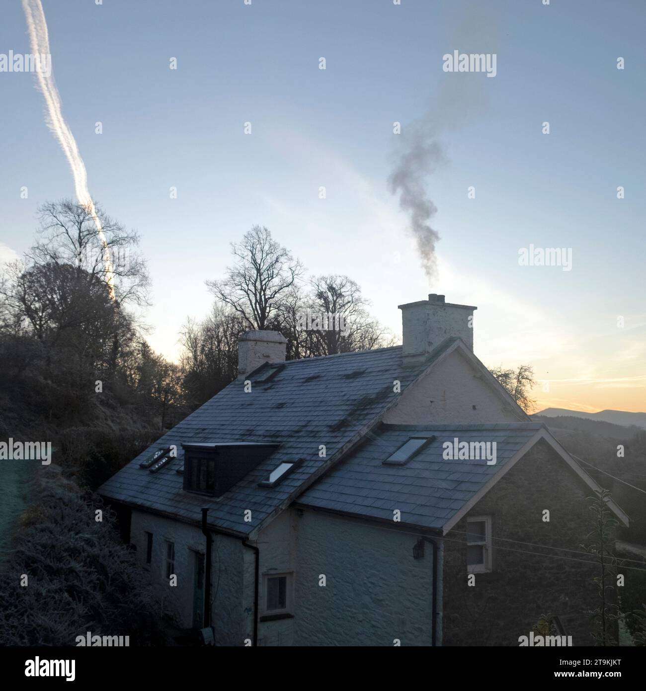 Fumo che si alza da una casa di campagna in inverno e tracce di vapore dall'aereo in inverno cielo blu rurale Galles UK KATHY DEWITT Foto Stock