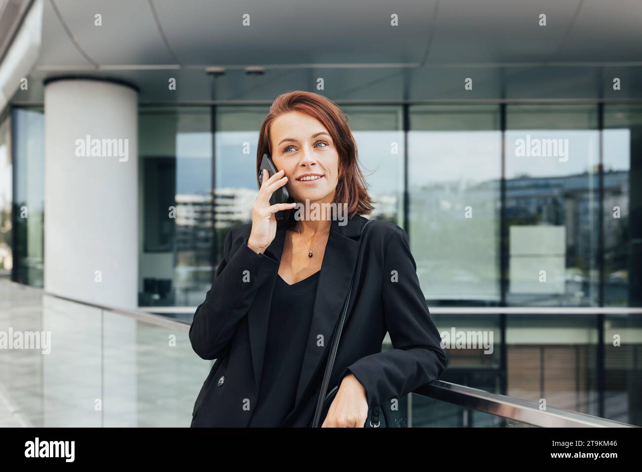 Donna d'affari sorridente di mezza età con i capelli di zenzero appoggiati su una ringhiera che parla su un telefono cellulare Foto Stock