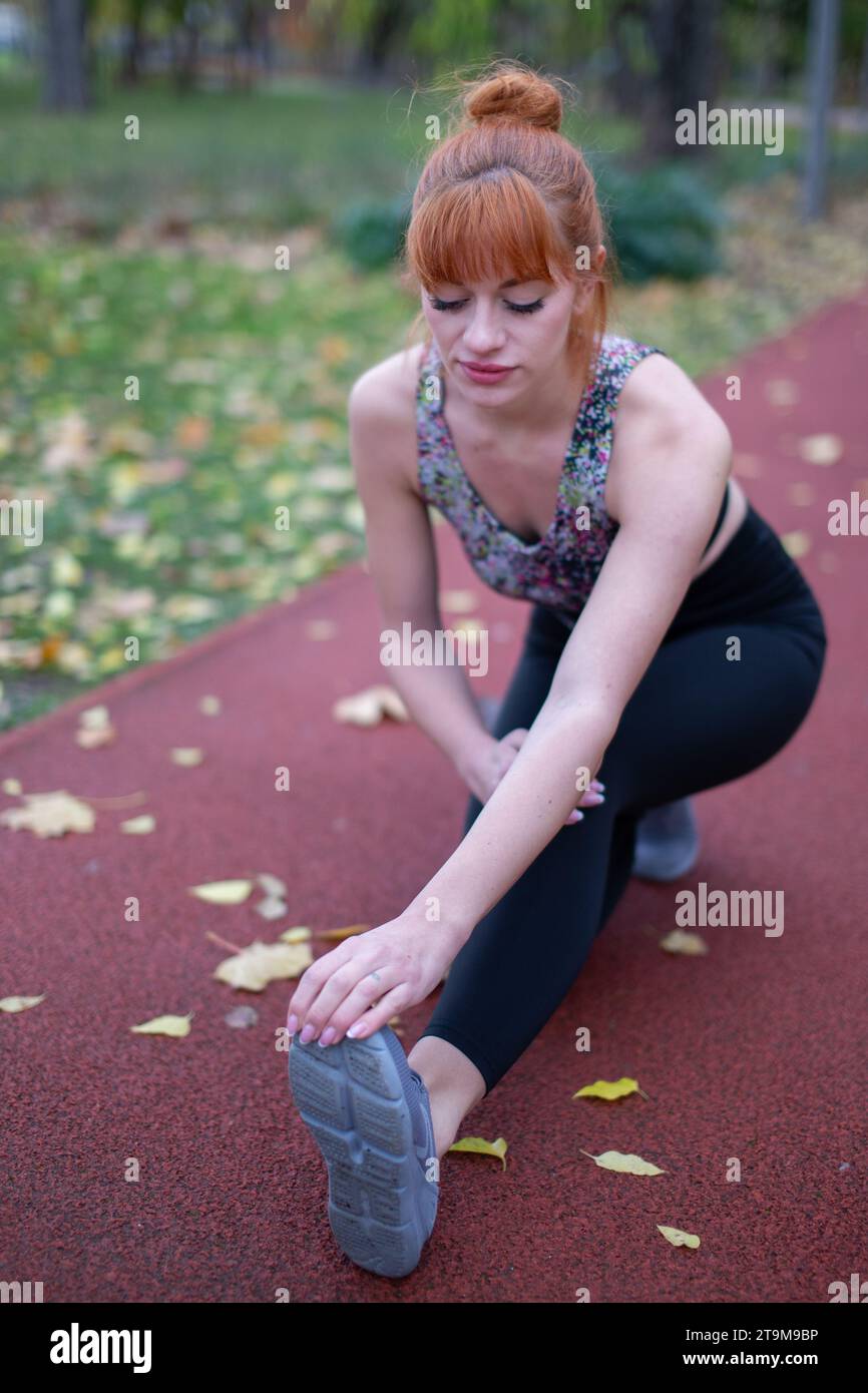 Giovane rossa donna caucasica gamba stretching si riscalda prima di correre in pista in autunno Foto Stock