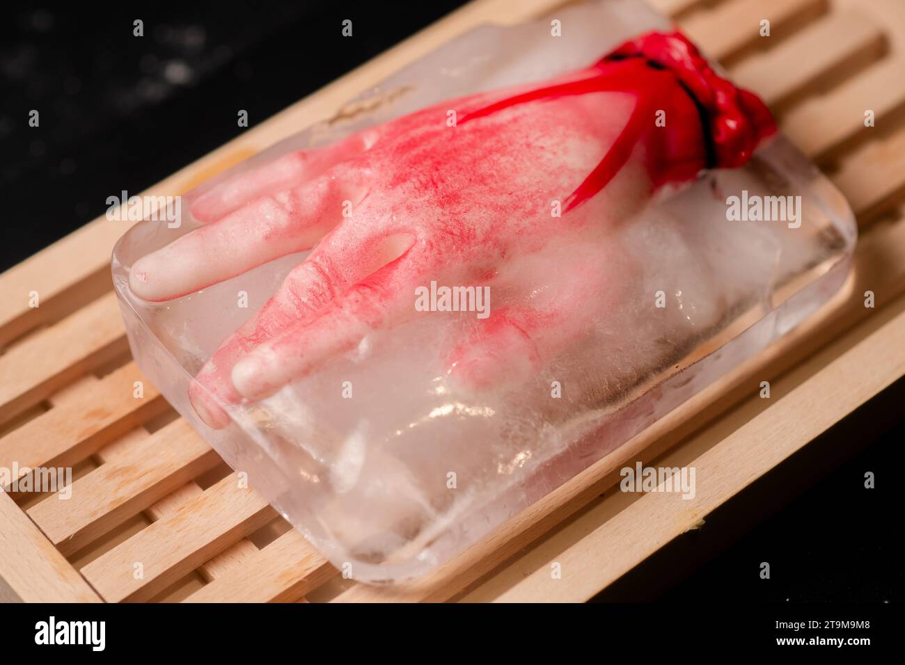 Orrore sul ghiaccio: Una finta mano umana congelata in un pezzo di ghiaccio su un tagliere di legno, creando una scena agghiacciante perfetta per i disegni a tema horror Foto Stock