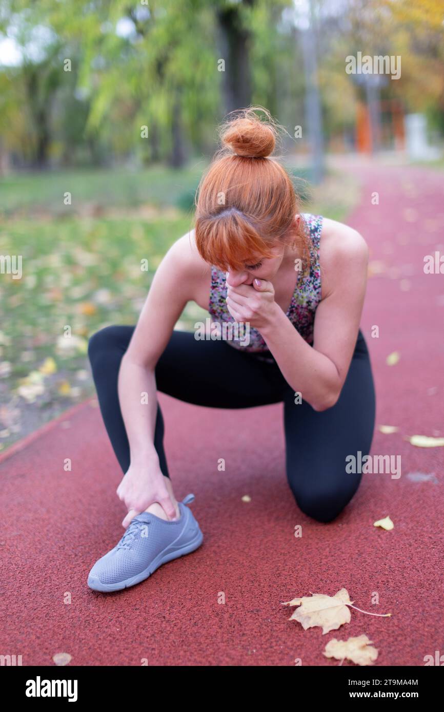 Donna caucasica Redhaed in abbigliamento sportivo con dolorosa lesione alla caviglia su pista da corsa Foto Stock