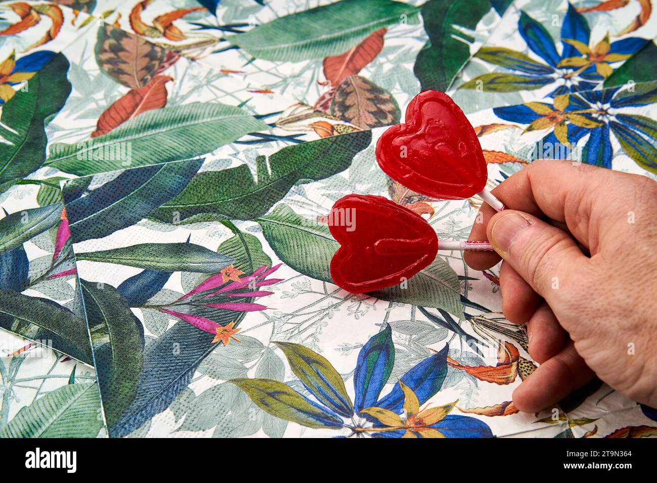 la mano dell'uomo regge due lecca lecca-lecca rossi a forma di cuore con uno sfondo tropicale. Foto Stock