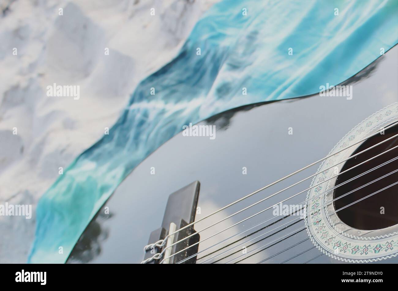Vista ravvicinata e prospettica di parte di una chitarra nera con pittura decorativa, adagiata su un sarong blu in stile tie-dye, sulla sabbia della spiaggia. Foto Stock