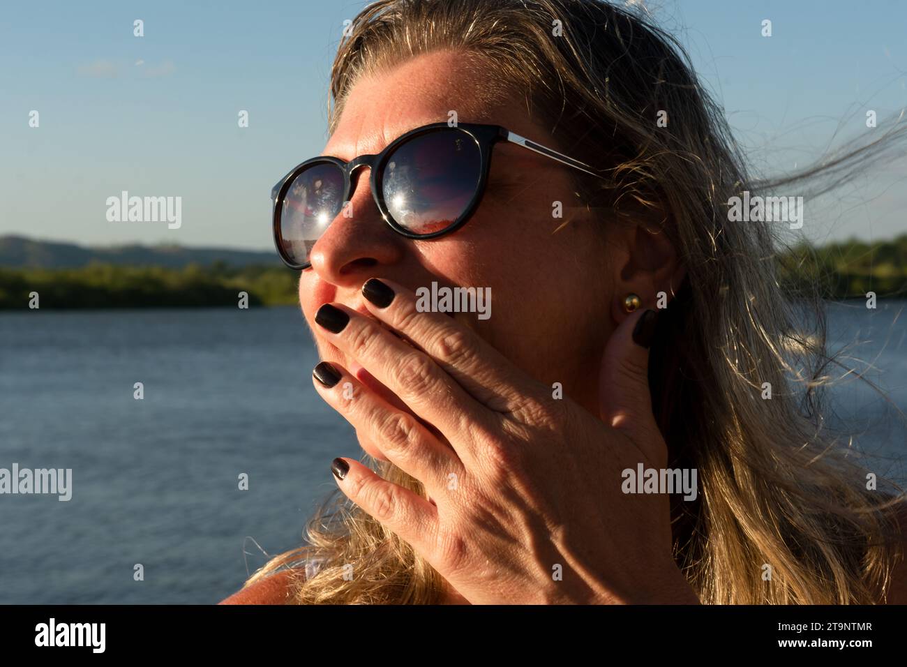 Ritratto del viso di una bella donna bionda che indossa occhiali da sole con la mano sulla bocca, contro un fiume. Foto Stock