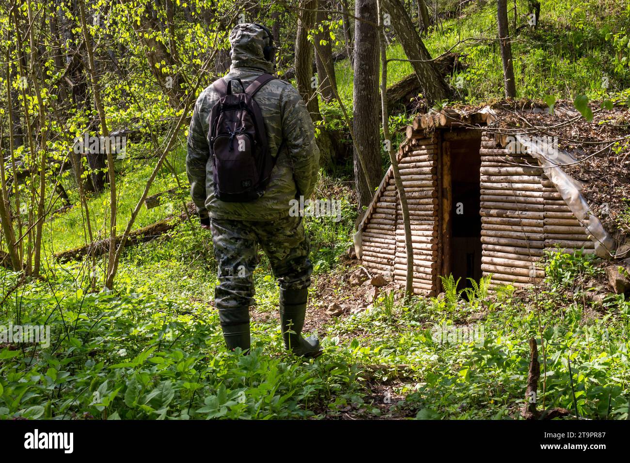 Un uomo con una tuta mimetica e uno zaino dalla schiena sullo sfondo di un dugout della foresta costruito in una foresta selvaggia Foto Stock