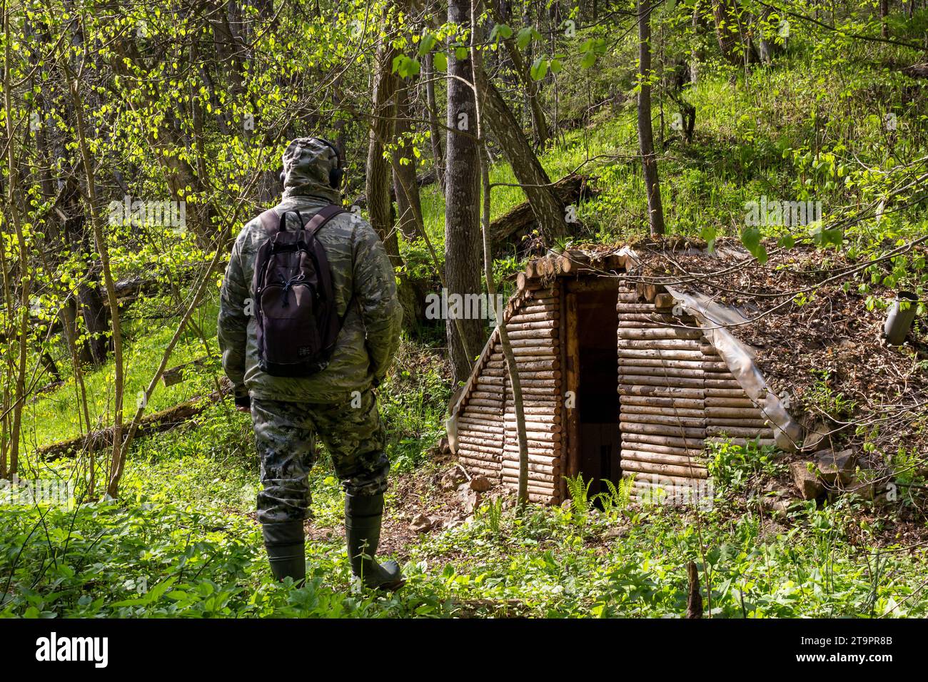 Un uomo con una tuta mimetica e uno zaino dalla schiena sullo sfondo di un dugout della foresta costruito in una foresta selvaggia Foto Stock