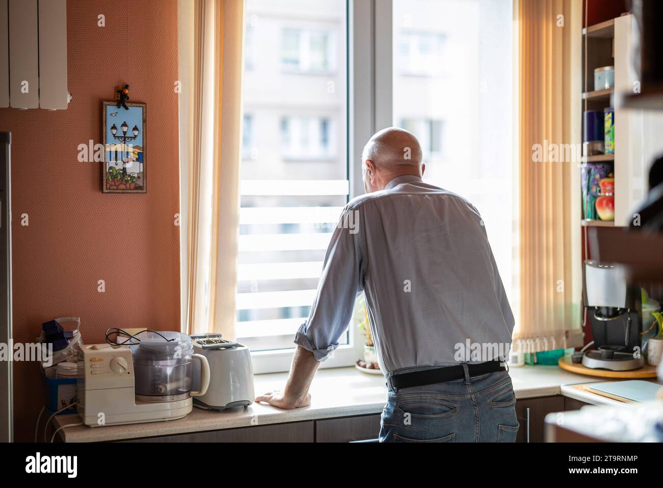 Uomo anziano in piedi al bancone della cucina in casa sua e guardando fuori dalla finestra Foto Stock
