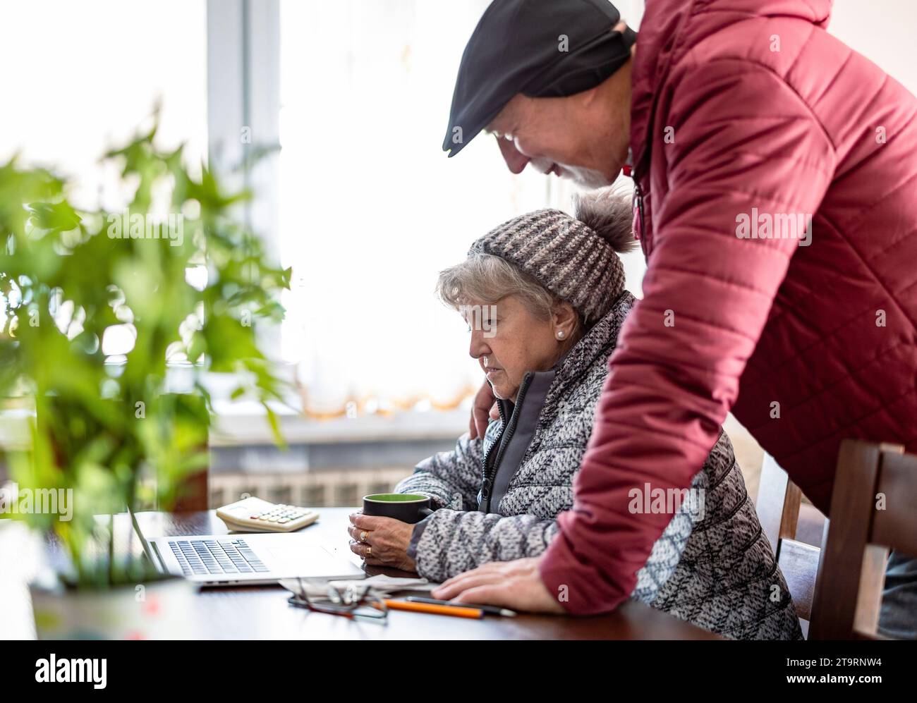 Coppia anziana che indossa abiti invernali che si occupa delle bollette energetiche sul notebook a casa Foto Stock