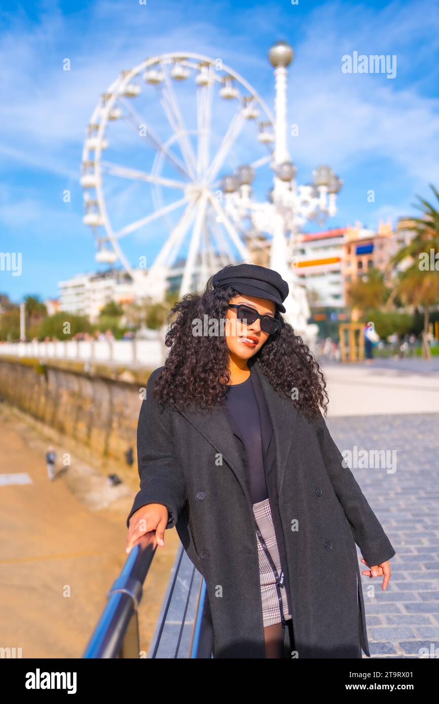 Ritratto di una donna latina alla moda in abiti invernali in piedi su una passeggiata accanto alla spiaggia in città Foto Stock