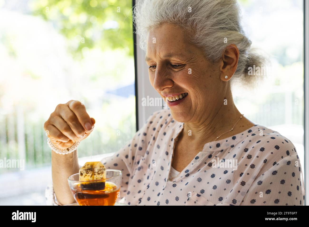 Una donna anziana caucasica felice che beve tè alla finestra nella soleggiata casa. Stile di vita, pensionamento, vita anziana e vita domestica, inalterati. Foto Stock