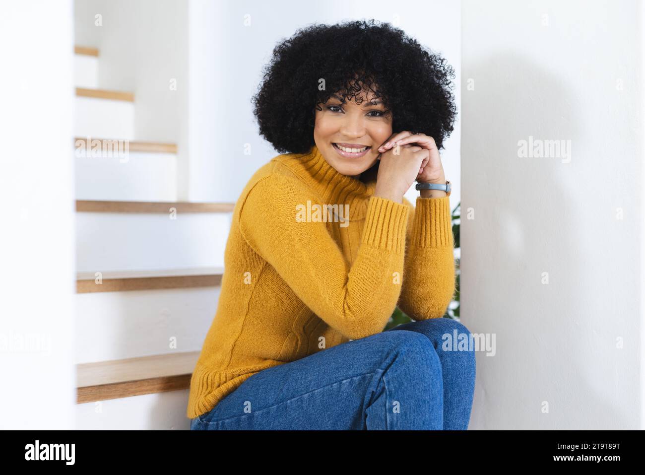 Felice donna afro-americana seduta sulle scale con le mani piegate a casa, spazio per le copie. All'interno, espressione, gesticolazione, linguaggio del corpo e femminilità, Foto Stock