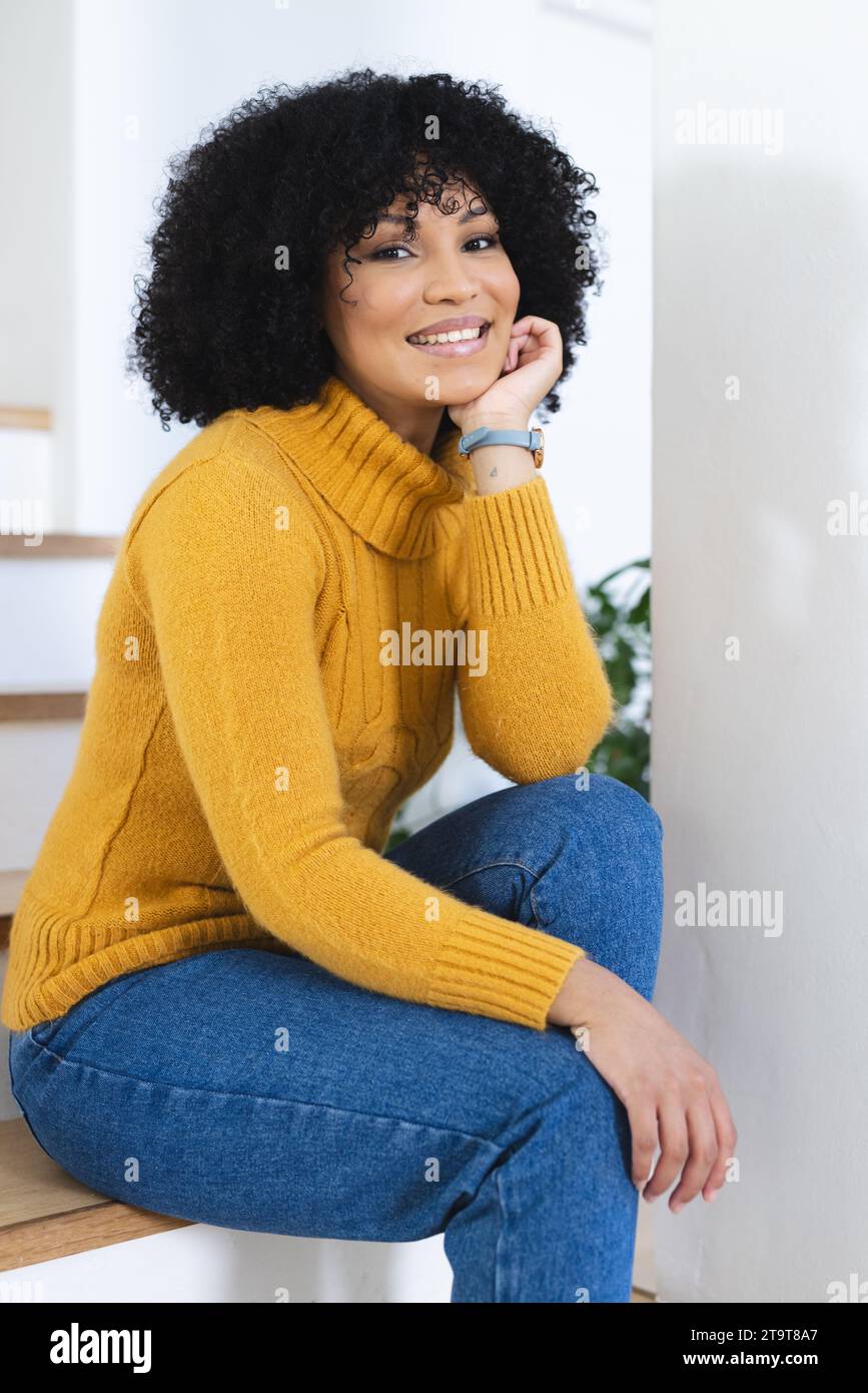 Felice donna afro-americana seduta con la mano sul viso sulle scale a casa, spazio fotocopie. All'interno, espressione, gesticolazione, linguaggio del corpo e femminilità, Foto Stock
