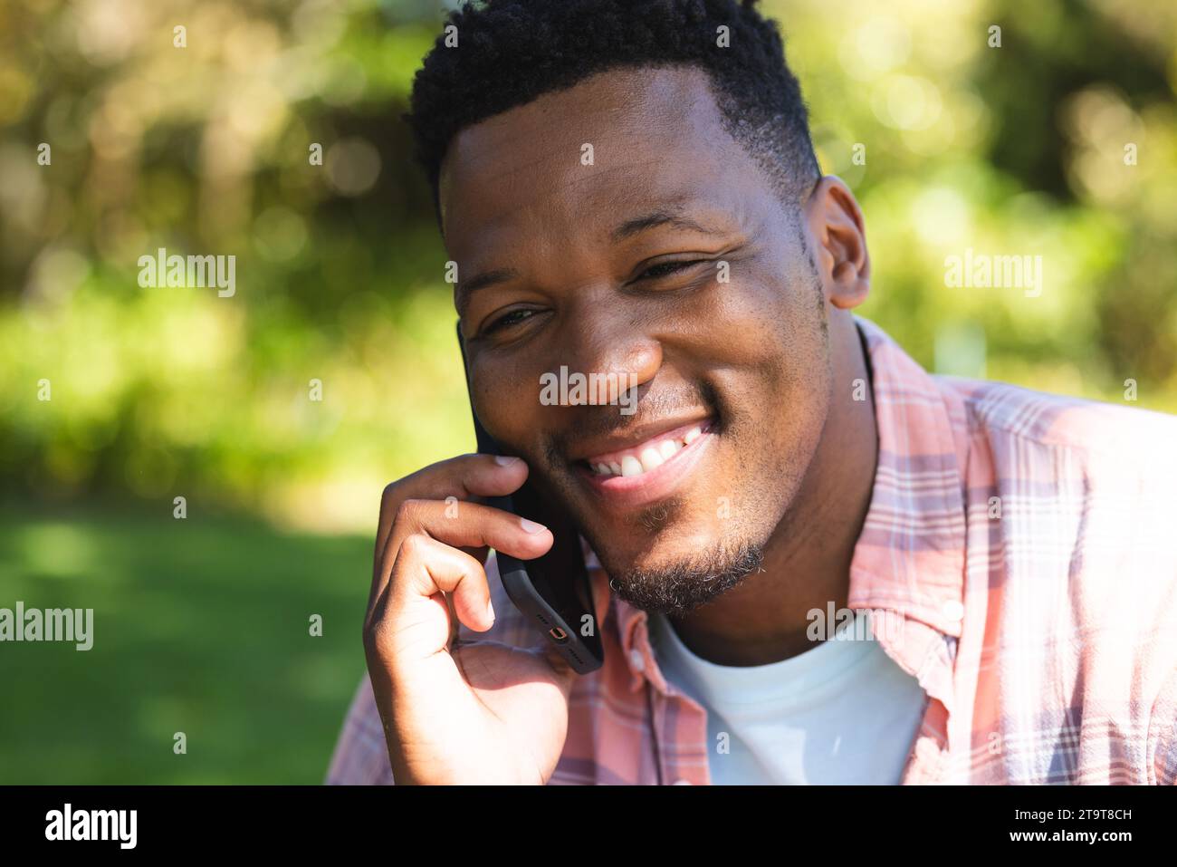 Felice afro-americano seduto sulle scale e parlando sullo smartphone nel giardino soleggiato, spazio fotocopie. Esterni, espressione, gesticolazione, tecnologia, c Foto Stock