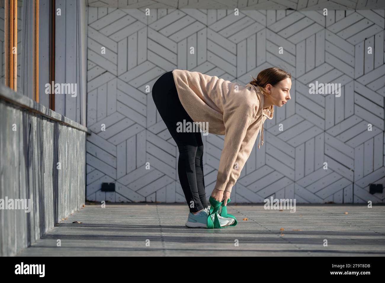 Una giovane atleta esegue esercizi di stretching per i muscoli della schiena e delle gambe con una cintura fitness, allenandosi con abbigliamento sportivo mentre si sta in piedi su una p di legno Foto Stock