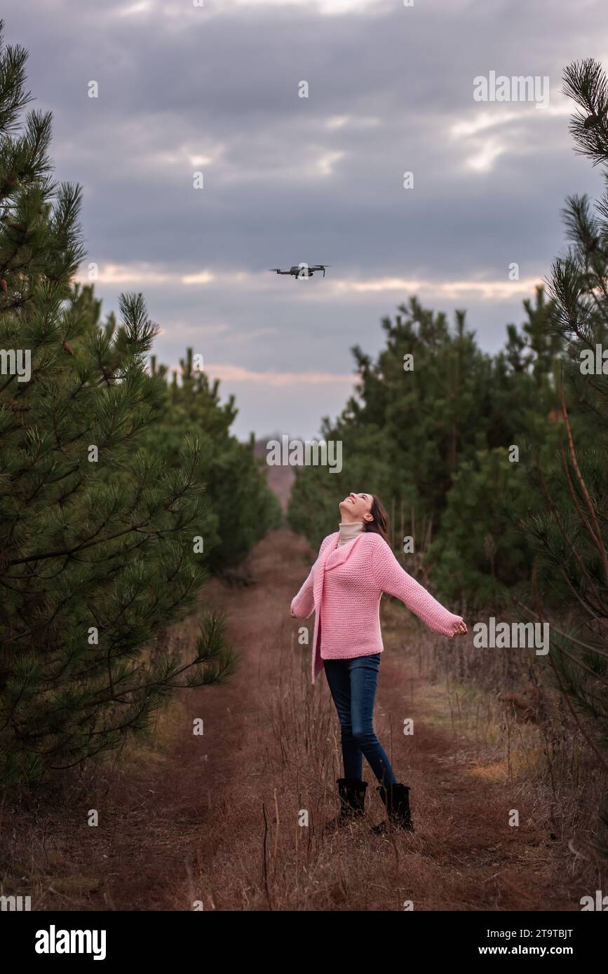 Giovane donna in maglia rosa in campagna tra alberi di conifere nella foresta in posa per una macchina fotografica drone nel cielo. Ragazza riccia che si diverte a nat Foto Stock